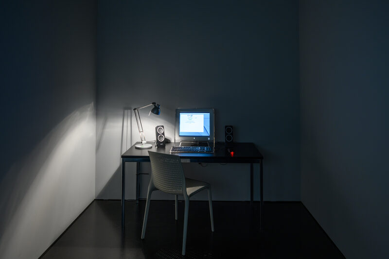 A period computer workstation installed in a darkened gallery: monitor, speakers, and desk lamp on a desk, lit only by the screen.
