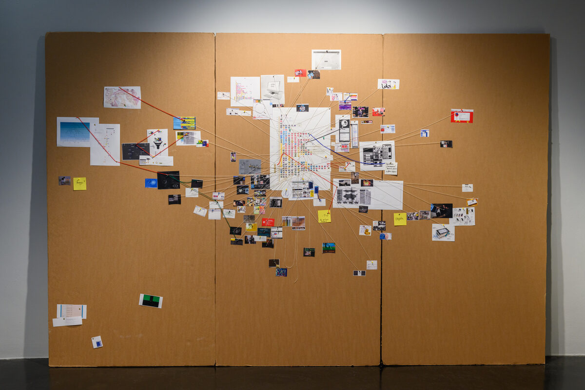 A wall-sized cardboard pinboard from the exhibition, covered in printouts, screenshots, and photographs joined by red and orange threads — the research behind the revivals, presented as a detective's evidence wall.